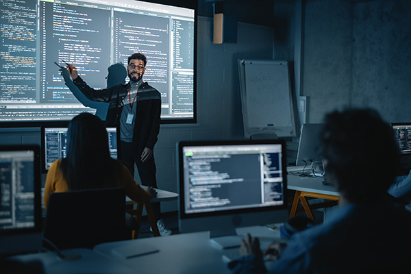 Teacher Giving Computer Science Lecture to Diverse Multiethnic Group of Female and Male Students in Dark College Room. Projecting Slideshow with Programming Code. Explaining Information Technology.
