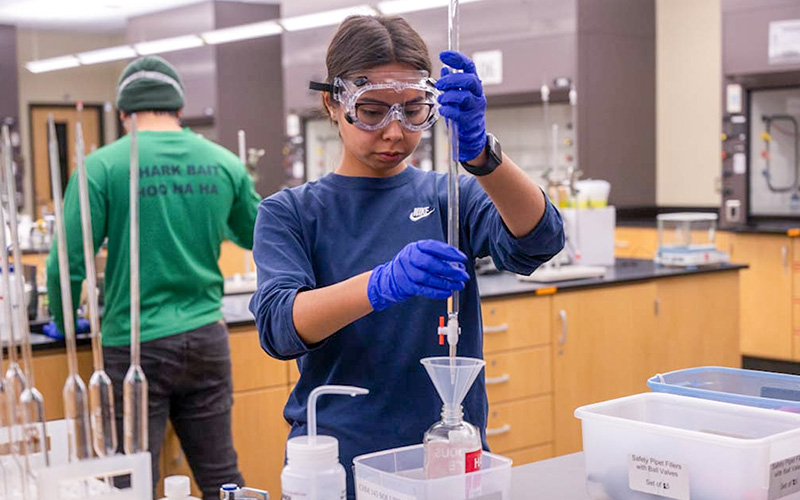 A female student, wearing safety glasses, is mixing chemicals in a glass flask bottle in the classroom.