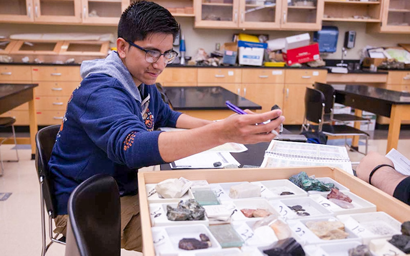 A male student is seated at a classroom table, examining various materials and taking notes.
