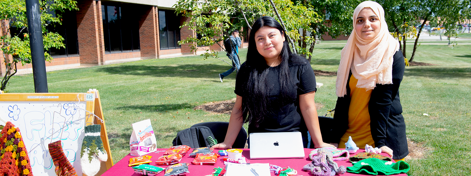 Two fiber arts club members display knitted and crocheted projects on a table.