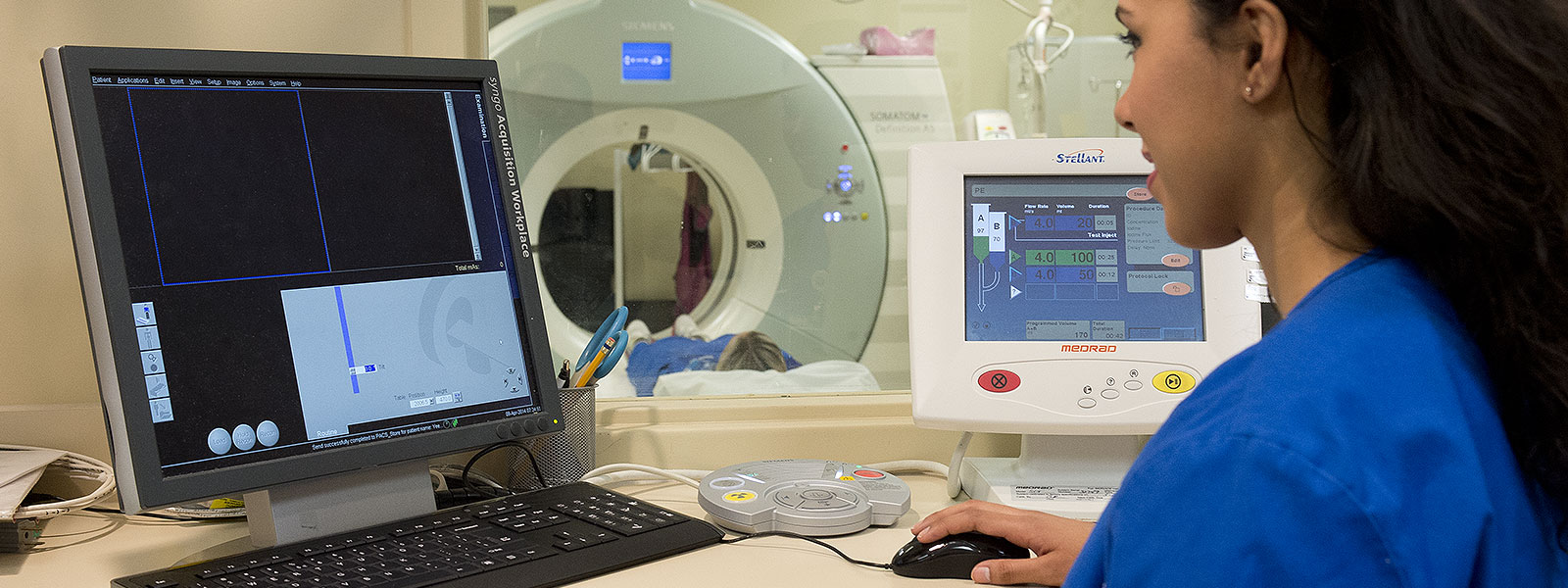 Close-up of medical technician working with imaging software on computer while patient is lying down on CT scanner in background.